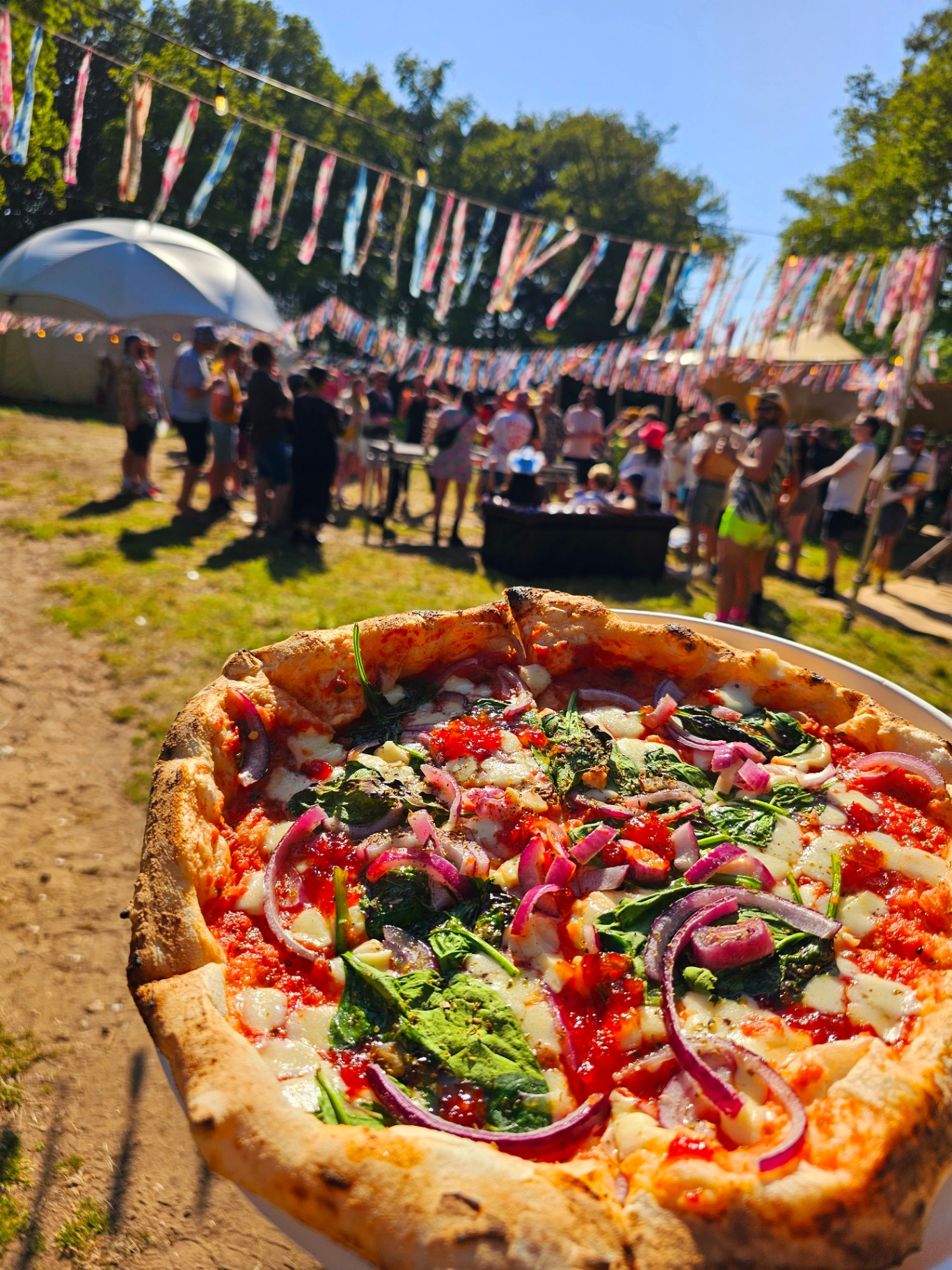 Wood-fired spinach and red onion pizza at a sunlit festival with bunting and crowd behind