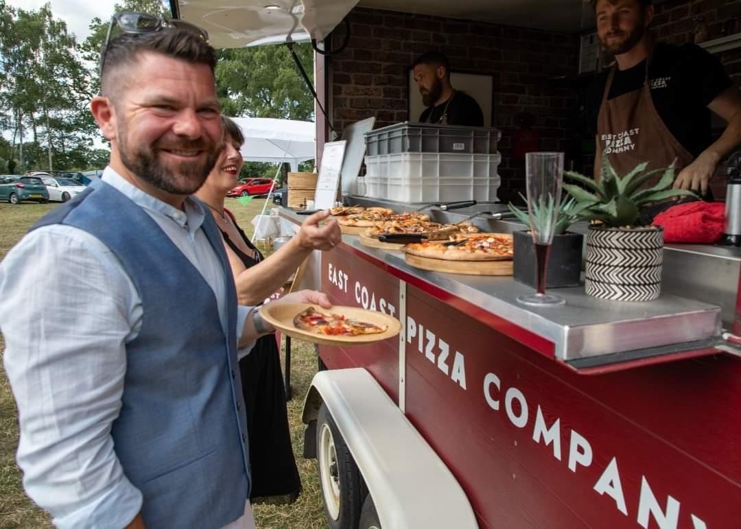 A smiling guest holding a pizza in front of the red Nag Box at a public event